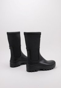 Bottes en caoutchouc noires avec une silhouette mi-mollet, finition texturée, bout rond et semelle extérieure épaisse et à motifs pour une bonne traction. Notez les panneaux latéraux.