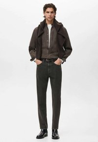 Prenda exterior de punto marrón sobre una camisa negra de botones, combinada con pantalones verde oliva y botas negras hasta el tobillo, en un fondo neutro.