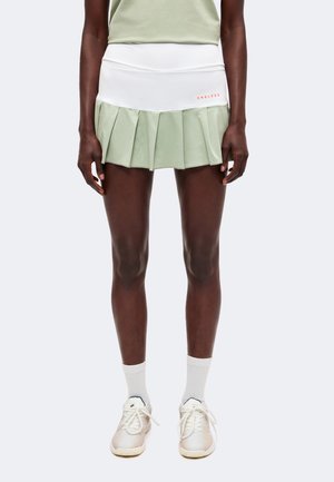 Persona che indossa una gonna da tennis plissettata bicolore bianca e verde chiaro con testo «"ENDLESS"», calzini bianchi e scarpe da ginnastica bianche, in piedi.
