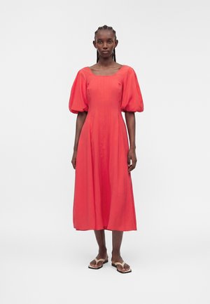 Femme aux cheveux tressés portant une robe midi corail avec des manches courtes bouffantes et un col carré, associée à des sandales plates beiges, debout.