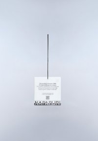 Etichetta bianca Napapijri con informazioni sulla sostenibilità riguardo al 100% cotone coltivato responsabilmente, appesa a un filo nero su uno sfondo chiaro.