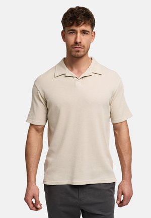 Jeune homme aux cheveux courts et bouclés portant un polo beige à manches courtes texturé et un pantalon foncé, sur un fond uni.