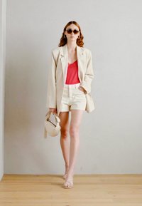 Model wearing a light beige oversized blazer, red lace camisole, white shorts, and nude strappy sandals, holding a small white bag.