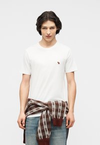 Νεαρός άνδρας με σκούρα μαλλιά φοράει λευκό T-shirt με μικρό λογότυπο ελάφου, μπλε τζιν και καφέ καρό πουλόβερ δεμένο γύρω από τη μέση.