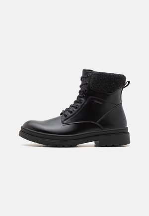 Bottine noire en cuir à lacets, avec col matelassé texturé et semelle épaisse en caoutchouc, vue de profil sur fond blanc.