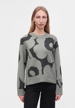 Jeune femme portant un pull gris avec de grands motifs floraux foncés et un pantalon noir, debout devant un fond clair uni.