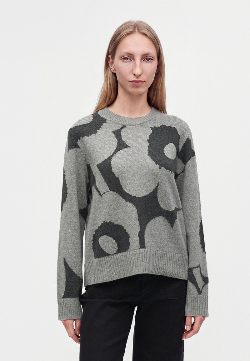 Jeune femme portant un pull gris avec de grands motifs floraux foncés et un pantalon noir, debout devant un fond clair uni.