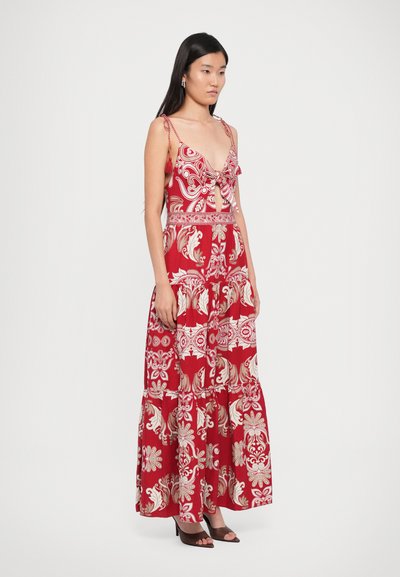 Maxi dress in red fabric with white floral and paisley patterns. Features a deep V-neck, bow detail, and tiered skirt. Worn with brown heels.