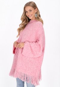 Pārmērīgs rozā adīts džemperis ar teksturētu apdari, zvana piedurknēm un frizētām malām. Valkāts ar ziliem džinsiem un lielām auskaru rotām.