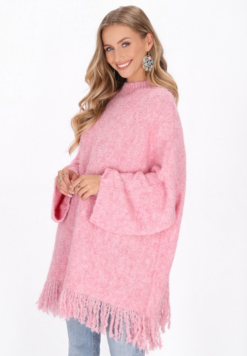 Pārmērīgs rozā adīts džemperis ar teksturētu apdari, zvana piedurknēm un frizētām malām. Valkāts ar ziliem džinsiem un lielām auskaru rotām.