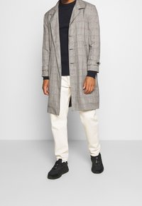 Grå rutigt kappa med snibbkrage, dubbelknäppt design och knappdetaljer, kombinerat med en marinblå tröja, krämfärgade byxor och svarta sneakers.