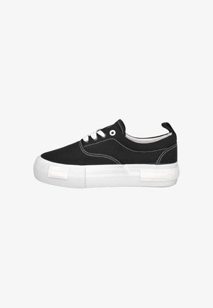Zapatilla negra de lona baja con suela de goma blanca, cordones blancos y costuras blancas sobre un fondo blanco liso.