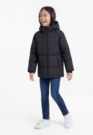 Smilende barn som har på seg en svart dunjakke med hette, blå jeans og hvite sneakers, står foran en ensfarget hvit bakgrunn.