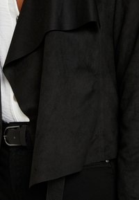 Veste en fausse suède noire avec un design devant drapé, présentant une texture lisse et un col à couches. Pressions visibles le long de la couture latérale.