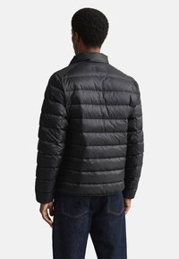 Juoda pūkinė striukė su horizontaliu šildymo raštu, stovinčiu apykakliu ir matinio paviršiaus. Dėvima ant tamsių džinsinių kelnių. Vaizdas iš nugaros.