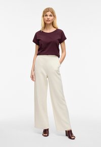 Bordo kratki top s kratkimi rokavi v kombinaciji s širokimi kremnimi hlačami z gladko teksturo. Rjave mule s peto dopolnjujejo obleko.