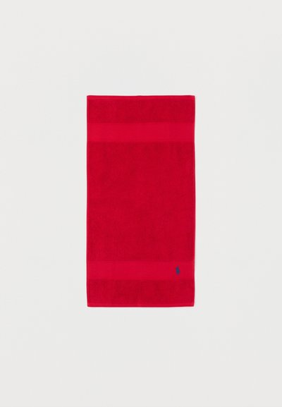 Rødt rektangulært håndklæde med to horisontale teksturerede striber og et lille mørkeblåt broderet logo nær den nederste kant på en hvid baggrund.