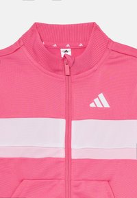 Rožinė zip-up striukė su raukšlėta apykakle, puošta baltomis horizontaliomis juostelėmis ir mažu baltu logotipu. Pagaminta iš minkšto, tekstūruoto audinio.