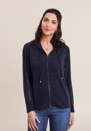 Haut à zip bleu marine avec cordons de serrage et détails en mesh sur les côtés, avec des manches longues et une coupe décontractée, associé à un jean bleu.