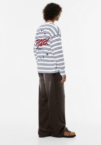 Langärmliges, weiß gestreiftes Hemd mit roter Grafik und Sternen auf dem Rücken, kombiniert mit braunen Cordhosen und braunen Schuhen mit blauen Akzenten.