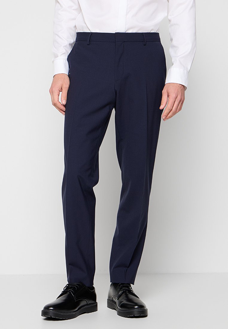 Mann in navyblauen maßgeschneiderten Anzughosen, weißem Hemd und schwarzen Leder-Schnürschuhen, der vor einem schlichten Hintergrund steht.