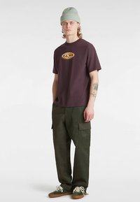 Ciemnofioletowa koszulka z okrągłym żółtym logo, w połączeniu z oliwkowozielonymi spodniami bojówkami z dużymi kieszeniami. Jasnozielona czapka beanie i sneakersy.