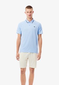 Ljusblå pikétröja med krage, med en mörk kant och en liten grön logotyp på bröstet. I kombination med beige shorts.