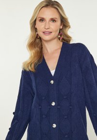 Námořnický modrý cardigan s diamantovým vzorem, texturovaná látka a velké stříbrné knoflíky. Má boční bambulové akcenty na rukávech.