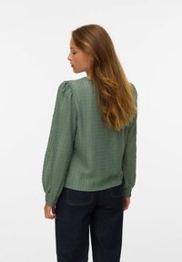 Femme aux longs cheveux bruns, portant un chemisier vert texturé à manches longues et un jean bleu foncé, de dos sur fond blanc.