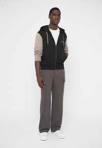 Sudadera negra con capucha y cremallera, mangas beige, combinada con pantalones grises de pierna ancha y zapatillas blancas. Tela suave y ajuste relajado.