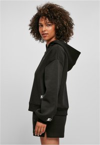 Sudadera con capucha negra de corte holgado, con puños acanalados y un pequeño parche de logo en el lateral. Combinada con pantalones cortos negros a juego.