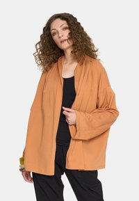 Pomarańczowy kardigan z długimi rękawami i luźnym krojem, wykonany z miękkiego materiału, z niezobowiązującym kołnierzem i otwartym przodem. Pod nim czarna bluzka.