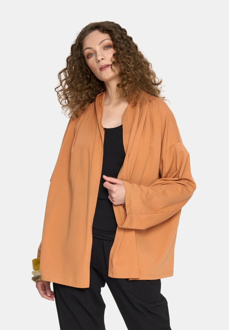 Pomarańczowy kardigan z długimi rękawami i luźnym krojem, wykonany z miękkiego materiału, z niezobowiązującym kołnierzem i otwartym przodem. Pod nim czarna bluzka.