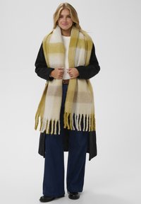 Donna che indossa un grande sciarpone a frange giallo e beige a quadri su un cappotto nero, un maglione bianco, jeans larghi blu e scarpe nere.