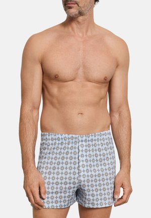 Homme torse nu portant un caleçon bleu clair à petit motif floral, debout devant un fond blanc uni.