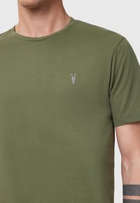 Oliivinvihreä puuvillapaita, jossa on pyöreä kaula-aukko. Vasemmalla rintakehässä on pieni harmaa brodeerattu pääkallo -kuvio. Pehmeä tekstuuri.