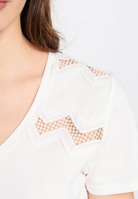 T-shirt bianca a V sul collo con dettagli in crochet a zigzag sulle spalle, che mette in risalto la texture e il design.