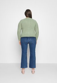 Pull vert à côtes avec des manches longues, associé à un jean évasé bleu et des sandales à talons roses pâles. Vue de dos montrant l'ajustement du vêtement.