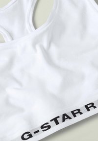 Bílá bavlněná sportovní podprsenka s dvojitým designem na zádech a černým logem "G-STARR" na spodní části. Hladká textura, jednoduchý styl.