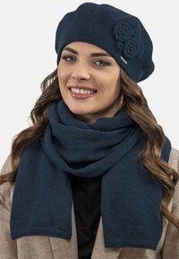 Béret en tricot bleu foncé avec trois accents floraux, associé à une écharpe assortie. Les deux articles ont une texture douce et confortable, ainsi qu'un design minimaliste.