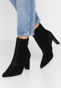 Bottines en daim noir avec bout pointu, talon bloc haut, et design à lacets comprenant des œillets et des lacets contrastants. Fermeture éclair sur le côté.