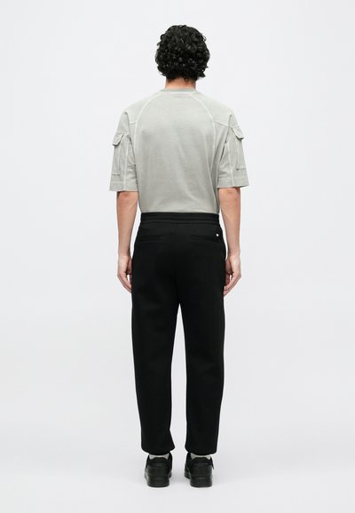 Šedá košeľa s krátkym rukávom a vreckami na ramenách, doplnená čiernymi nohavicami. Oblečenie má voľný strih a textúru látky.