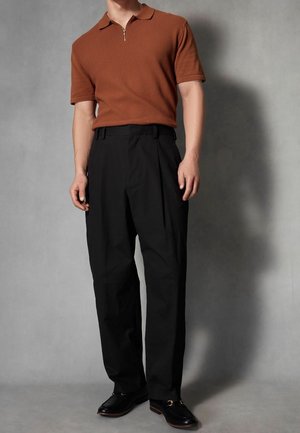 Polo marron à manches courtes et fermeture éclair sur le devant avec un détail texturé, associé à un pantalon noir plissé et des mocassins en cuir noir avec des détails en or.