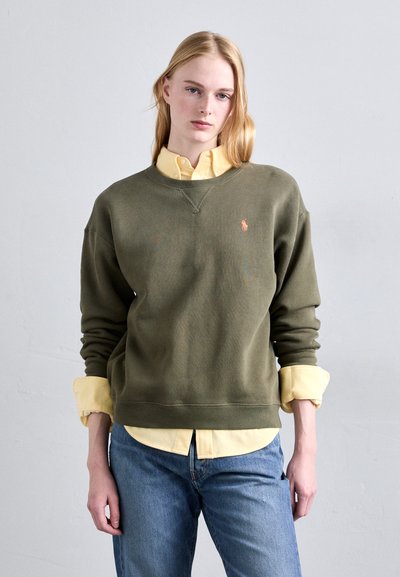 Sweatshirt vert olive avec des poignets et un ourlet côtelés, arborant un petit logo orange. Superposé à une chemise bouton éclair jaune clair et un jean bleu.