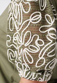 Olijfgroene blouse met witte geborduurde bloemenontwerpen; heeft een textuur met doorzichtige mouwen en een gemaakte manchetdetail.