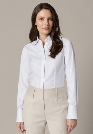 Kobieta o długich brązowych włosach, ubrana w dopasowaną białą koszulę zapinaną na guziki i beżowe spodnie z wysokim stanem na neutralnym tle.