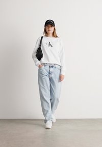 Jeune femme marchant à l'intérieur, portant un sweat-shirt blanc Calvin Klein, un jean bleu clair, des baskets blanches, une casquette noire et portant un sac à bandoulière noir.