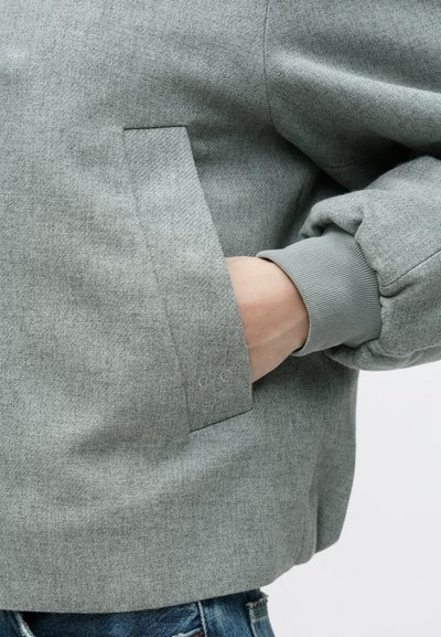 Veste gris clair en tissu texturé, dotée d'une poche latérale, d'un poignet élastique et d'une coupe décontractée. Détails de couture visibles.