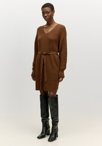 Braun gestricktes Kleid mit V-Ausschnitt, tailliert mit einem Gürtel. Lange Ärmel mit gerippter Textur. Kombiniert mit knielangen schwarzen Stiefeln.