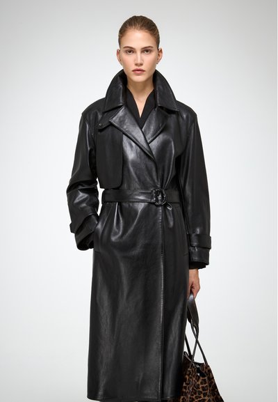 Donna con capelli tirati indietro indossa un trench coat in pelle nera con cintura e tiene una borsa con stampa leopardata su uno sfondo semplice.